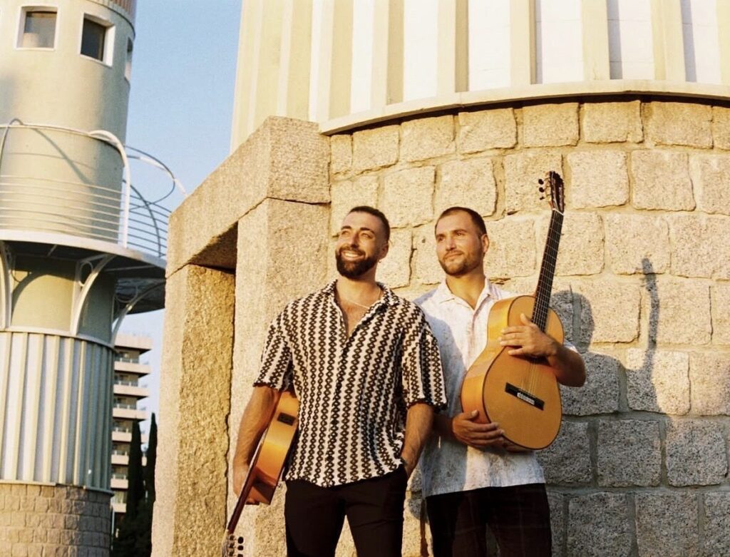 Zwei Musiker posieren mit Gitarren neben einer Steinmauer.