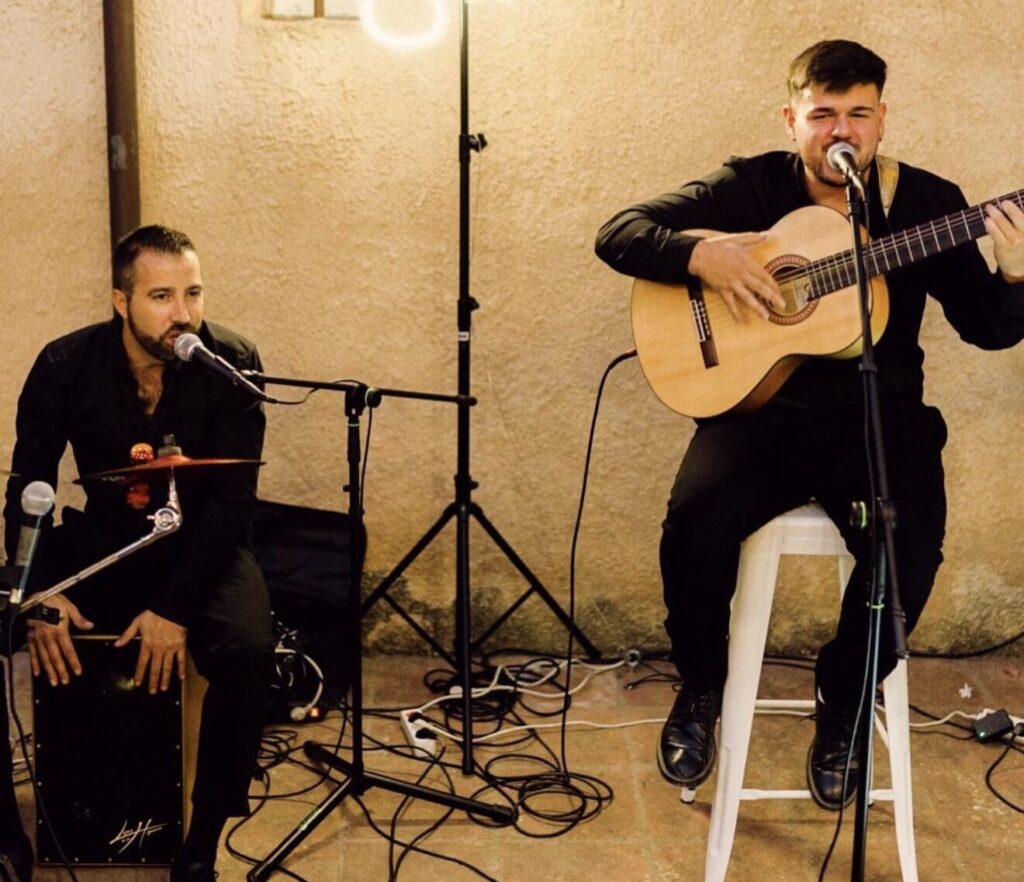 Guitarrista y percusionista sentados, cantando ante sus micrófonos.