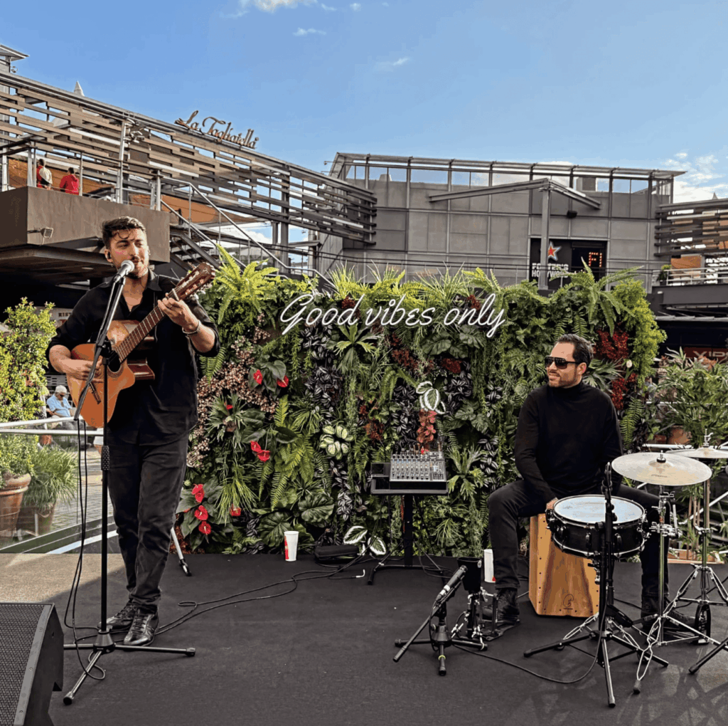 Guitarrista y percusionista actuando junto a un fondo vegetal.