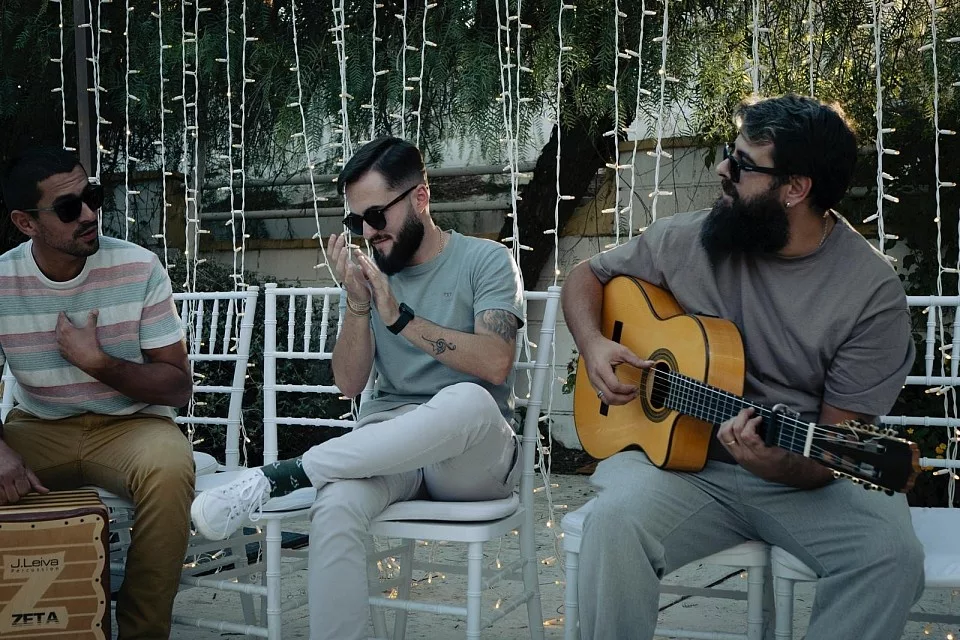 Tres músicos con guitarra, palmas y cajón sentados al aire libre.