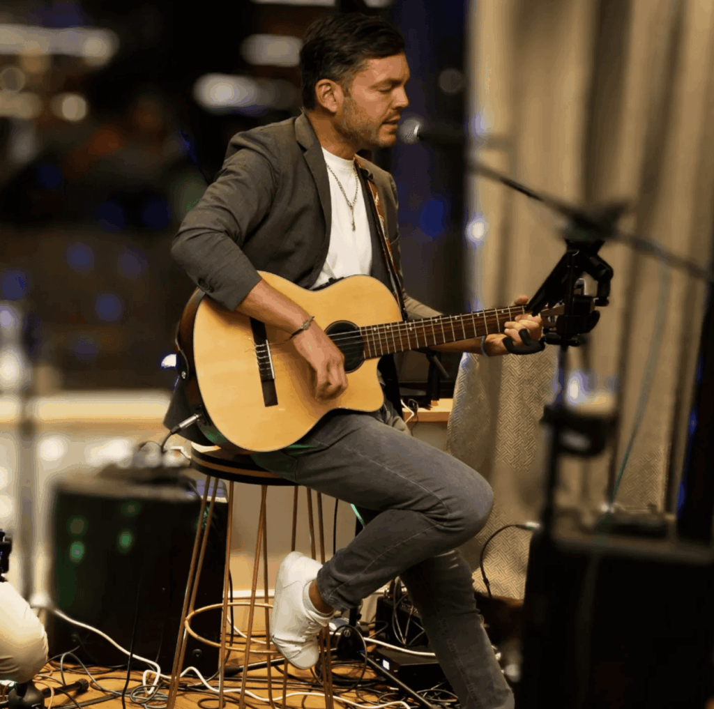 Músico sentado cantando y tocando la guitarra ante un micrófono.